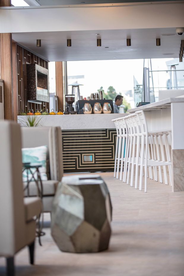 A bright, refined contemporary commercial bar and lounge interior. The space features a long white bar with a row of white bar stools, a geometric black-and-brass decorative bar front, pale flooring, integrated ceiling spotlights, timber wall panelling, and a textured dark feature wall behind the counter. Lounge seating and a sculptural faceted side table occupy the foreground, while large windows provide an airy backdrop.