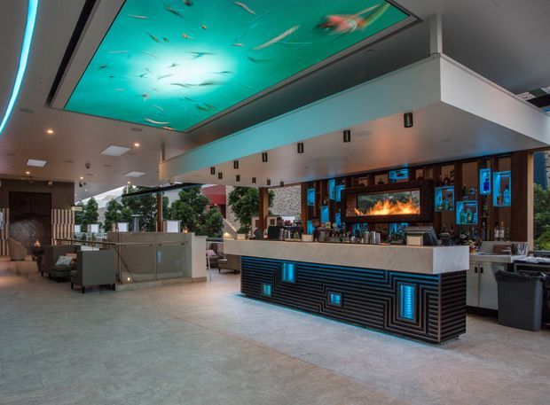 A spacious contemporary commercial interior featuring a dramatic illuminated ceiling panel depicting underwater fish, a large bar with a geometric black ribbed facade and glowing blue inset details, and a back bar with a fireplace-like illuminated feature. Pale stone or concrete-look flooring, dark timber cabinetry, open glazing, and lounge seating create an upscale hospitality atmosphere.