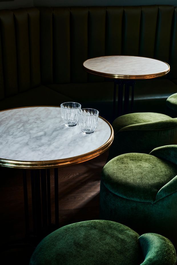 Moody close-up of an upscale lounge or dining area featuring round white marble-topped tables with polished brass edging and dark slender bases. Deep green velvet upholstered chairs and banquette seating surround the tables, with two faceted drinking glasses on the foreground table. The space has dramatic low lighting, dark tones, and a luxurious, intimate atmosphere.
