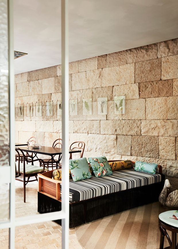 A warm, eclectic living and dining area viewed through a glazed or mirrored partition. The room features a substantial rough beige stone-block feature wall, a black-and-white striped daybed or sofa with colourful botanical cushions, a mid-century-style wooden armchair, and a dark dining table with bentwood chairs. Light timber or terracotta-toned flooring, framed botanical artwork, and soft natural light create a layered, relaxed interior.