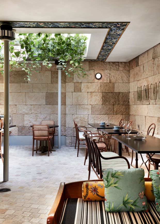 A refined indoor-outdoor dining space with rough rectangular stone walls, pale patterned stone or ceramic paving, dark dining tables, and curved timber-and-cane chairs. A large skylight or open roof section brings in daylight and supports trailing greenery, while a decorative dark floral border frames the ceiling opening. The foreground includes a striped upholstered lounge with colourful botanical cushions, creating an eclectic restaurant-like atmosphere.