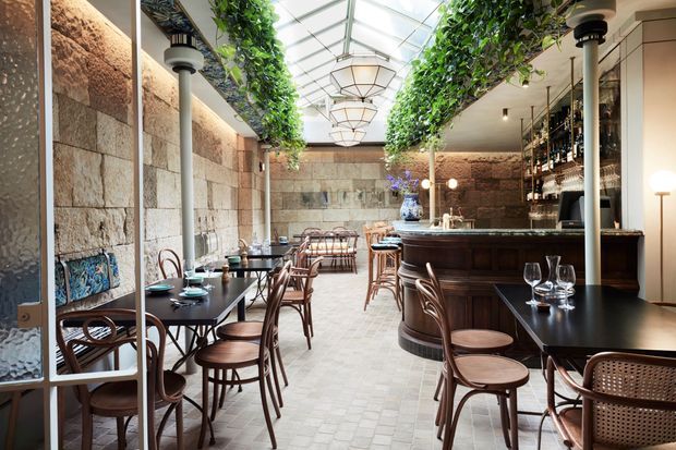 A refined restaurant or wine bar interior with exposed textured stone walls, dark timber bar joinery, black dining tables, bentwood and cane chairs, and a glazed atrium roof. Hanging greenery runs along the ceiling edges, while decorative pendant lights and a backlit bottle display create a warm, elegant atmosphere.