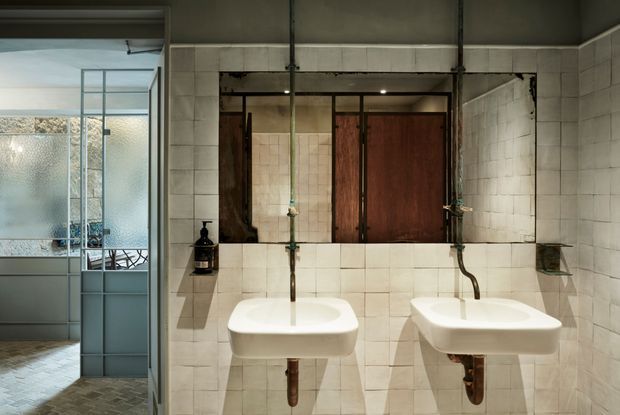 Industrial-style bathroom with two wall-mounted white ceramic basins, exposed plumbing, aged metal wall-mounted taps, and a large timber-framed mirror cabinet. The walls are clad in small pale square tiles, with dark concrete-like ceiling surfaces and utilitarian detailing. A frosted-glass partition and glimpse of an adjacent shower or wet area are visible at left.