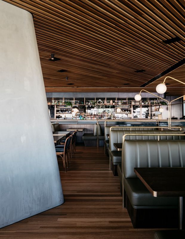 A sophisticated restaurant and bar interior viewed from the dining area, with dark timber flooring, a slatted timber ceiling, upholstered booth seating, dining tables, a long back bar with extensive bottle shelving, and sculptural globe pendant lights. A large curved pale plaster feature wall frames the left side of the space.