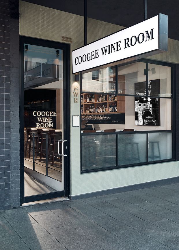 Street-facing exterior of the Coogee Wine Room, showing a dark-framed glazed entrance and shopfront, illuminated signage, pale small-format tiled facade, and a visible wine bar interior with bottle shelving, bar seating, and artwork.