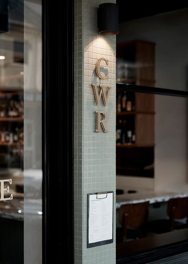 A close-up architectural detail of a hospitality or commercial interior entrance, featuring a pale green small-format tiled column with raised metallic letters spelling “CWR,” a black cylindrical wall light, black-framed glazing, and a dimly visible bar or dining area beyond. The space has a moody, refined atmosphere with dark timber joinery and black accents.