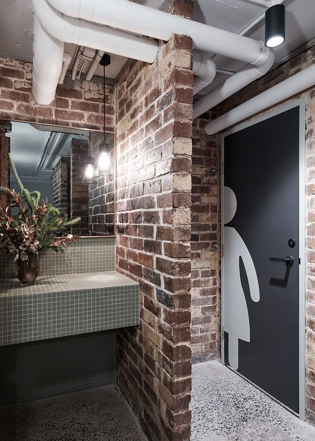 A compact industrial-style interior with exposed red brick walls, visible white-painted services and ductwork across the ceiling, dark framed glazing, and a black graphic door. A tiled vanity with integrated basin sits beside a large mirror, while terrazzo-style flooring and pendant lighting complete the utilitarian fit-out.
