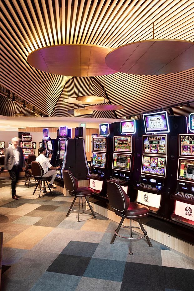 Interior of a casino or gaming venue with rows of electronic gaming machines, upholstered swivel stools, patterned carpet, and a dramatic slatted timber ceiling with large suspended oval acoustic or decorative panels. Warm ambient lighting and dark gaming-machine finishes create a lively commercial atmosphere.