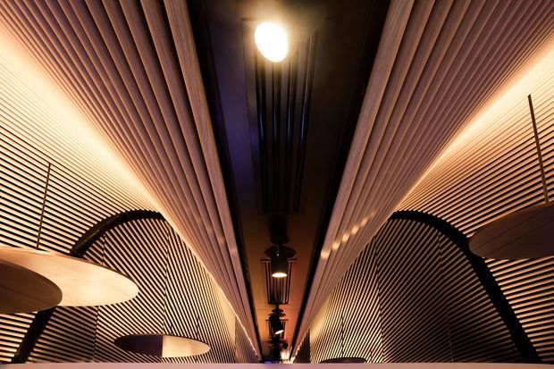 Dramatic interior architectural view of a narrow, elongated ceiling or passage-like space featuring extensive warm timber slatted wall and ceiling lining. Curved black structural arches, suspended pendant lights, and a dark central ceiling recess create a rhythmic, high-contrast contemporary composition.