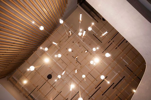 Upward-looking view of a tall interior ceiling featuring a suspended grid ceiling with numerous hanging globe and linear pendant lights. A curved white architectural edge frames the right side, while closely spaced timber slats create a textured ceiling feature along the left.