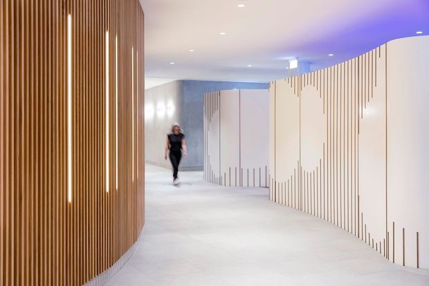 A contemporary interior corridor or lobby with sweeping curved walls. The left wall is clad in closely spaced vertical timber slats with integrated linear lighting, while the right side features large pale panels detailed with irregular vertical grooves. Recessed ceiling lights and blue-purple cove lighting create a polished, futuristic atmosphere. A person is visible in the distance, emphasizing the scale of the space.