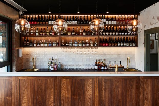 A refined bar interior with a long timber-clad front counter, white tiled backsplash, dark ceiling, brass fixtures, and extensive open shelving displaying bottles and hanging stemware. Four spherical pendant lights provide warm illumination, creating a sophisticated hospitality-style atmosphere.