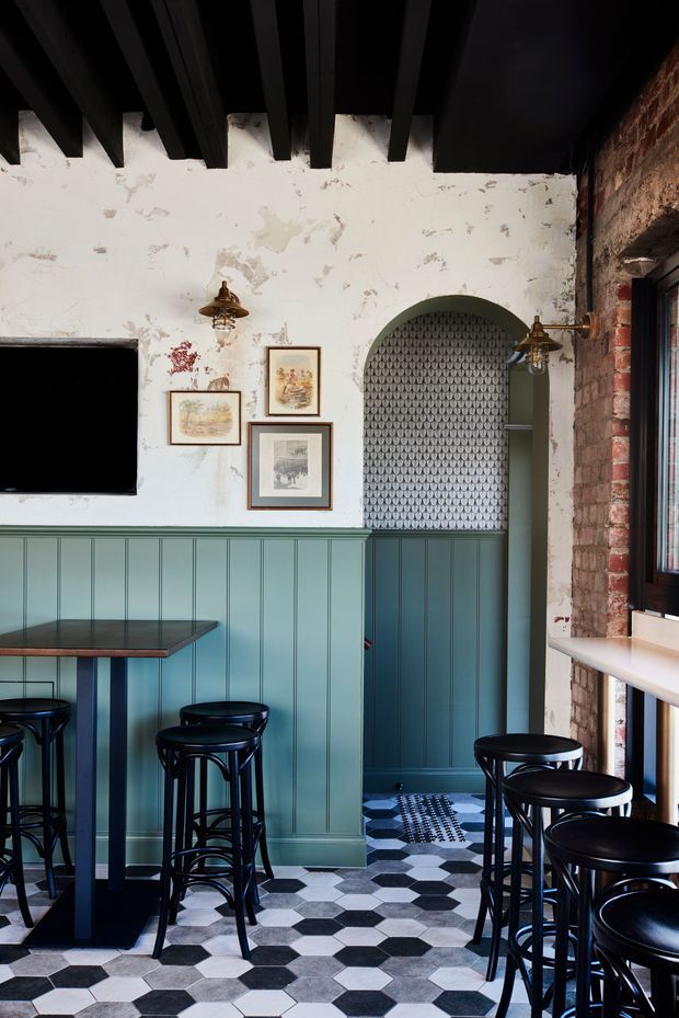 A compact, moody bar or café interior with a teal-blue panelled half-wall, patterned wallpaper in an arched alcove, exposed red brick, black-painted exposed ceiling beams, and a bold black-and-white geometric tiled floor. Black bar stools and narrow tables line the space, with vintage-style brass wall lights and framed artwork adding character.
