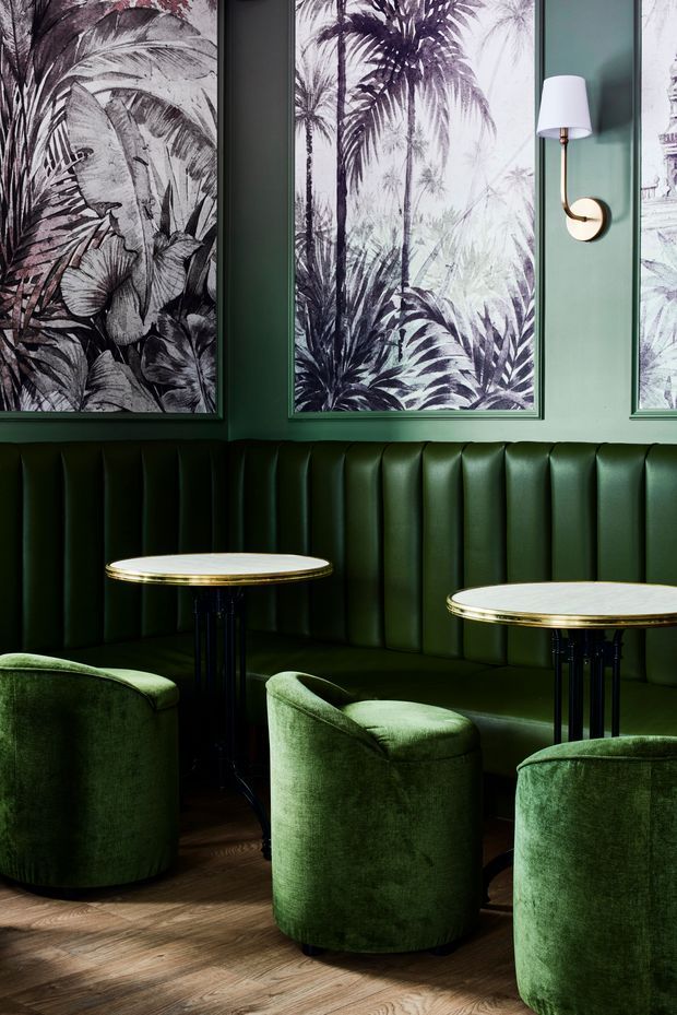 A refined hospitality-style seating nook with deep forest-green walls and vertically channelled upholstered banquette seating. Two small round marble-topped tables with dark slender bases sit among rounded green velvet armchairs. Botanical monochrome artwork in dark frames, brass wall lighting, and warm timber flooring create a moody, luxurious atmosphere.
