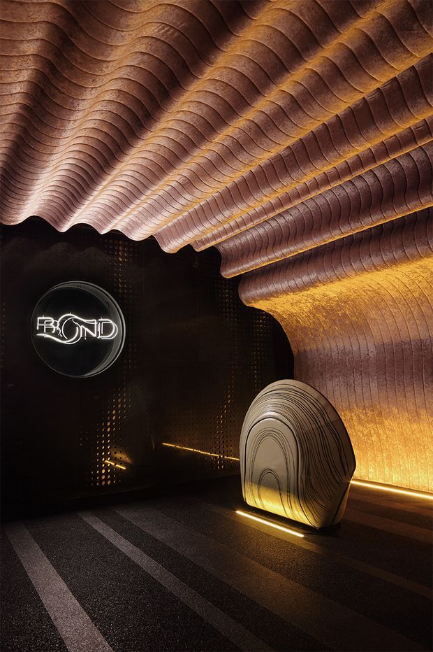 A dramatic dark commercial interior or branded venue featuring a sculptural undulating ceiling and wall installation in layered copper, bronze, and warm brown tones. Concealed amber lighting traces the organic forms and illuminates a large sculptural object near the wall. The space includes a circular illuminated sign, dark patterned walls, and a carpeted floor with linear tonal bands. The overall effect is immersive, luxurious, and highly sculptural.