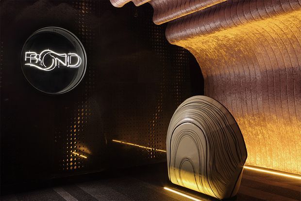 Moody, highly stylised interior detail featuring a glowing “BEYOND” sign on a dark perforated wall, warm concealed linear lighting, sculptural layered wood-like forms, and a large organic rounded feature element. The space has a dramatic hospitality or entertainment atmosphere with strong contrast between black surfaces and amber illumination.