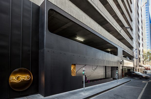 Urban exterior view of a multi-storey commercial building, dominated by a dark metal-clad street frontage and a large recessed, open-sided structure resembling a parking or service entrance. Concrete upper levels, narrow glazing, bollards, pavement, and adjacent high-rise buildings reinforce the dense city setting. A circular illuminated 'BOND' sign is mounted on the facade.