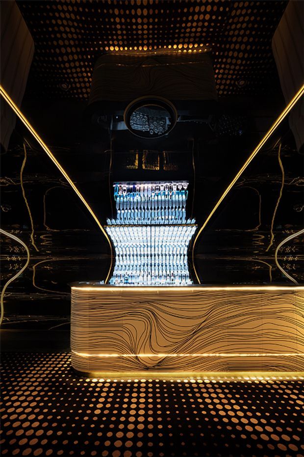 A dramatic, highly stylised commercial bar or reception counter in a dark interior. The foreground features a curved, illuminated counter with flowing wood-grain patterned cladding, backed by a glowing display of bottles and a central reflective circular feature. Black glossy wall surfaces, warm gold linear lighting, and a perforated patterned ceiling and floor create a luxurious, futuristic atmosphere.