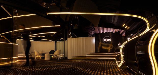 Dark, dramatic commercial interior with glossy black reflective surfaces, illuminated curved linear light installations, ribbed partitions, patterned flooring, and a raised stage or feature wall displaying a logo. The space has a futuristic, immersive showroom or entertainment-venue atmosphere.