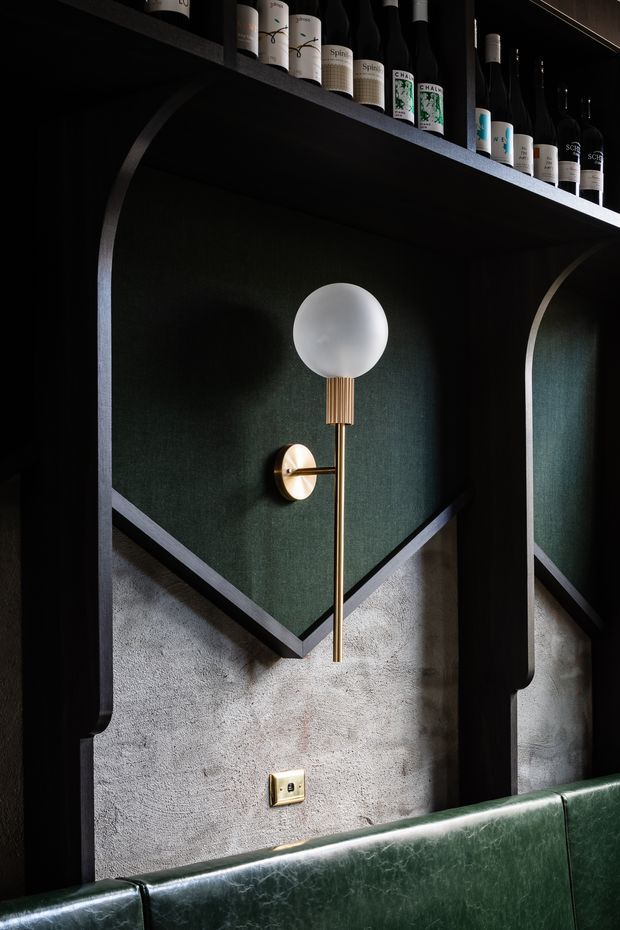 A dark, richly detailed interior feature wall with deep green upholstered or textured inset panels framed by black architectural joinery and curved vertical edges. A brass wall sconce with a white globe shade is mounted centrally above a green leather banquette. Open shelving along the top displays product bottles, suggesting a refined hospitality, bar, salon, or commercial interior. The lower wall has a rough grey textured finish, with brass electrical fittings adding a warm accent.