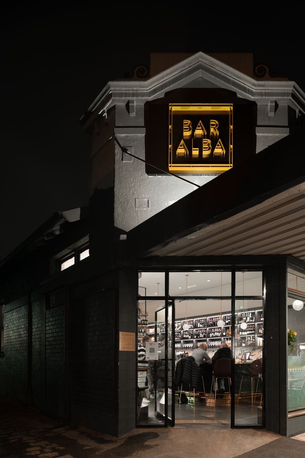Night-time exterior view of a bar housed in a renovated-looking masonry building. A prominent illuminated yellow BAR sign sits beneath a decorative classical gable, while large black-framed glazed doors and windows reveal the illuminated bar interior, bottle displays, and patrons. Dark brick walls, a covered entrance canopy, and wet paving create a dramatic streetscape atmosphere.