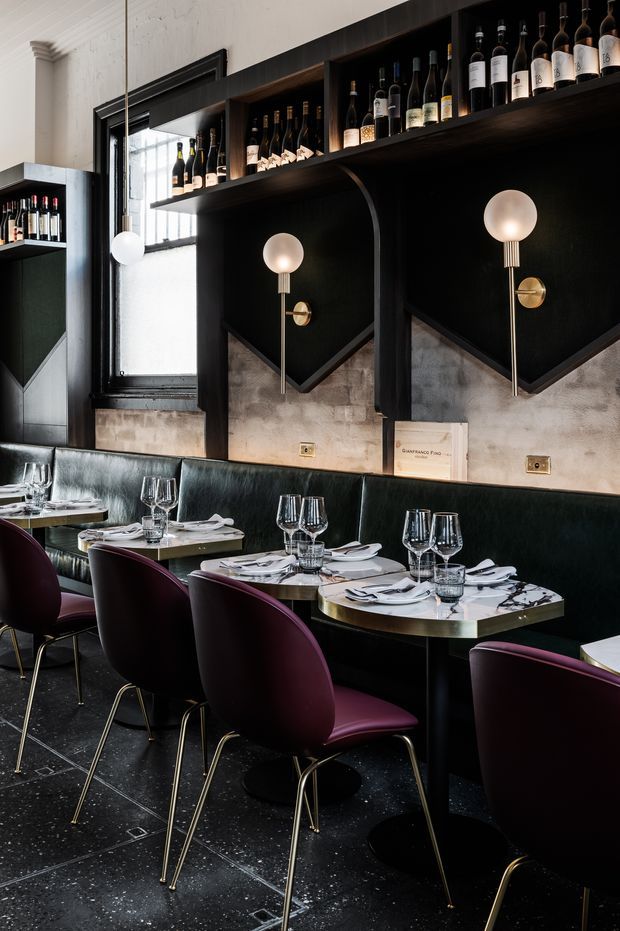 A refined restaurant dining area with a long dark banquette, burgundy upholstered dining chairs, marble-topped tables, and black terrazzo-style flooring. Dark timber or painted shelving displays wine bottles above sculptural black wall panels, with globe wall sconces providing warm lighting. The space combines dramatic dark finishes with brass details and light stone surfaces.