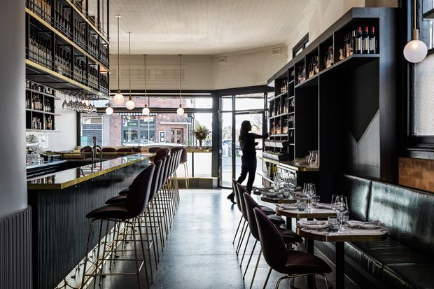 A refined contemporary restaurant and wine bar interior with a long central bar, dark ribbed joinery, extensive wine shelving, upholstered bar stools, booth seating, and set dining tables. Large glazed openings bring in daylight and frame an urban streetscape. Pendant globe lights, brass-toned detailing, and a white timber-lined ceiling contrast with the dark cabinetry and leather seating.