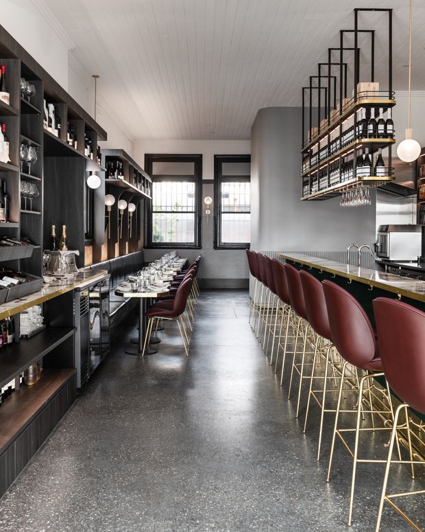 A refined commercial bar and dining interior with a long counter lined by burgundy upholstered bar stools with brass-toned legs. Dark built-in shelving displays bottles and glassware along the left wall, while suspended bottle racks hang above the bar. A banquette dining area with small tables occupies the rear, framed by tall black-trimmed windows. The space features a white beadboard ceiling, dark polished flooring, grey walls, and spherical pendant lights, creating a sophisticated hospitality setting.