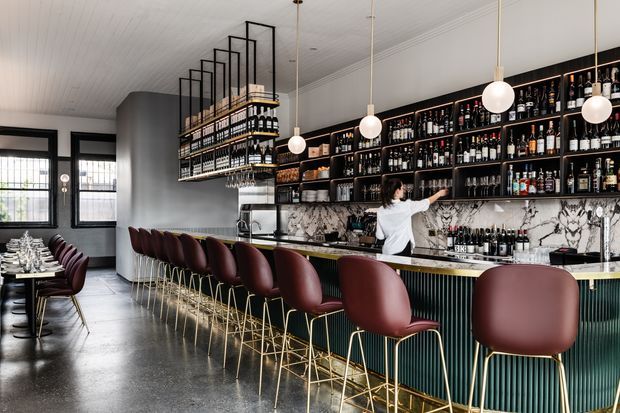 A stylish commercial bar and dining interior with a long dark green fluted bar front, white marble-look splashback, extensive black open shelving filled with bottles and glassware, and a suspended overhead bottle display. Burgundy upholstered bar stools with slim gold-toned frames line the counter, while spherical pendant lights hang from a white panelled ceiling. A dining area with set tables is visible to the left, framed by black industrial-style windows.