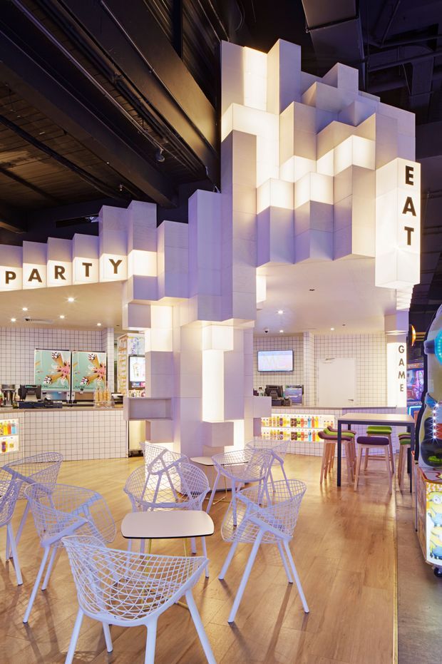 A brightly lit commercial entertainment and dining space with a playful geometric interior. The room features a large sculptural installation of stacked white illuminated blocks, tiled service counters, colourful signage reading “PARTY” and “EAT,” white wire lounge chairs and tables, timber-look flooring, and a bar-height seating area. The exposed dark ceiling and visible services create a strong contrast with the glowing white forms.