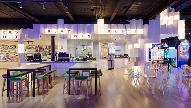 A brightly lit, retro-styled indoor arcade and entertainment space with multiple arcade machines, game counters, high communal tables, stools, and café-style seating. The room features a dark exposed industrial ceiling, warm wood-look flooring, tiled service counters, and a dramatic suspended geometric white feature installation with integrated lighting.