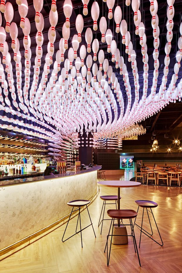 A striking commercial bar and dining venue with a long illuminated bar counter, extensive bottle display, timber herringbone flooring, dark timber ceiling and wall finishes, and rows of wooden dining tables in the background. The ceiling is covered with a dramatic installation of suspended bowling pins, while small round tables and tall stools occupy the foreground. Warm ambient lighting and brass-toned details create an energetic, theatrical hospitality interior.