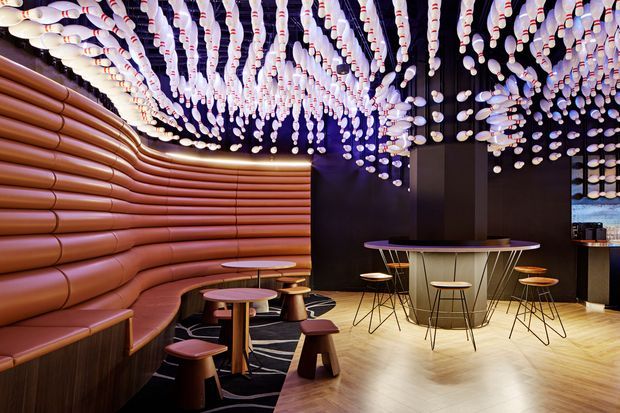 A dramatic contemporary hospitality interior, likely a bar or lounge, featuring a curved upholstered banquette with horizontal terracotta-brown ribbing along the left wall, small round tables, stools, and a central high communal table. The dark ceiling is covered with an elaborate installation of suspended white and pale pink bulb-like elements, creating a striking canopy above the warm timber-toned floor. Dark walls and ambient purple lighting add contrast and a theatrical atmosphere.