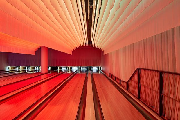 A dramatic, symmetrical interior view of a bowling alley with multiple polished wooden lanes extending toward the pins. The space is illuminated in saturated red and orange light, with a sculptural ribbed ceiling canopy, dark red wall treatments, and a metal safety railing along the right side.