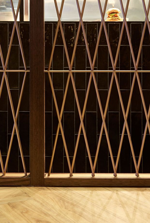 Close-up interior architectural detail featuring a dark glossy tiled wall behind a decorative geometric metal screen with repeated diamond-shaped diagonals. A substantial dark timber vertical mullion and base rail frame the screen, while pale timber flooring runs across the foreground. Warm lighting and a small burger display visible near the upper right suggest a refined commercial hospitality interior, likely a restaurant or café.