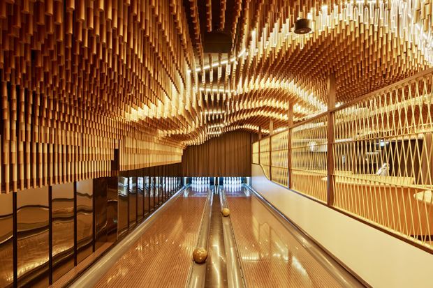 A dramatic bowling alley interior with highly reflective lanes, dark gutters, and glowing amber-toned timber slat detailing covering the ceiling and walls. Repeated suspended wood elements create a sculptural, wave-like canopy, while geometric metal or timber screening lines the right side. The space has a polished, immersive and luxurious commercial entertainment aesthetic.