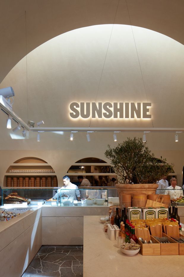 A warmly lit contemporary food-service interior, likely a café or restaurant, viewed from the customer side of a long stone counter. The space features a dramatic curved cream ceiling with a large illuminated “SUNSHINE” sign, arched wall openings with built-in shelving, a glass-fronted food display, track lighting, an olive tree in a large planter, and organised countertop merchandise and serving items. Staff and food preparation areas are visible behind the counter.