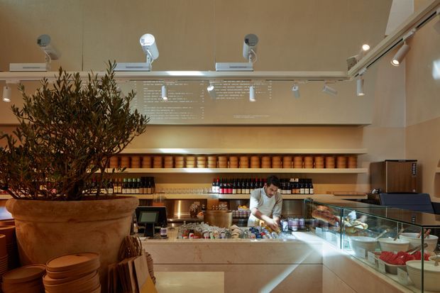 A warmly lit contemporary commercial food-service interior, likely a bakery, café, or deli counter. The image shows a long pale stone or concrete service counter with a glass display case containing prepared food, a worker behind the counter, extensive open shelving stocked with jars and bottles, track lighting, and projected or printed menu text on the rear wall. A large potted tree and stacked wooden serving ware add natural and tactile elements.
