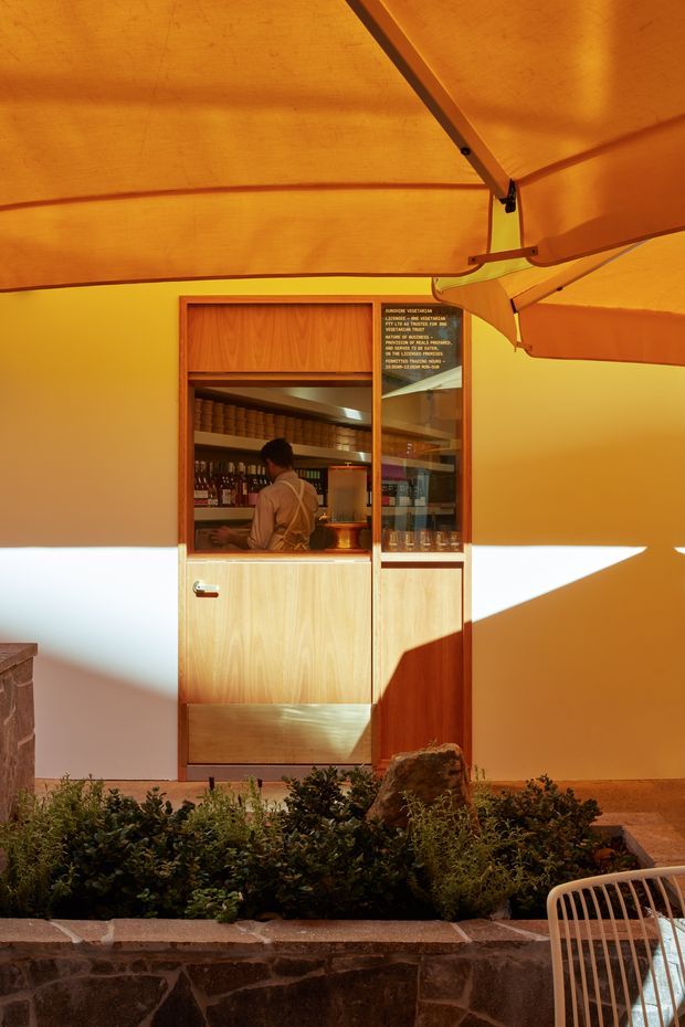 A warm, amber-lit service kiosk or bar built into a tent-like outdoor enclosure. The compact timber-framed serving window has glass shelving stocked with bottles, jars and glasses, with a staff member visible behind it. An orange fabric canopy and structural supports span overhead, while a landscaped planter with low greenery and a rough stone retaining wall occupies the foreground. The scene has a distinctive temporary hospitality or event-space character.