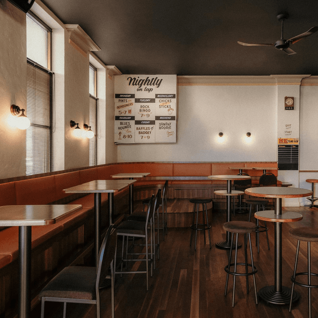A warmly lit retro-style café or casual dining space with a long rust-coloured upholstered banquette, small round and rectangular tables, dark metal chairs and stools, timber flooring, cream plaster walls, tall windows with blinds, decorative wall sconces, and a dark ceiling with a ceiling fan. Vintage menu boards and typography reinforce the nostalgic diner atmosphere.