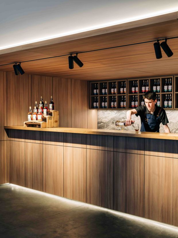 A sophisticated built-in bar counter with a bartender serving drinks. The space is clad extensively in warm vertical timber panelling and cabinetry, with open bottle shelving, a pale stone or marble-look backsplash, a timber benchtop, dark inset counter detailing, concealed under-counter lighting, and black track spotlights set into the timber ceiling. The atmosphere is warm, refined, and hospitality-oriented.