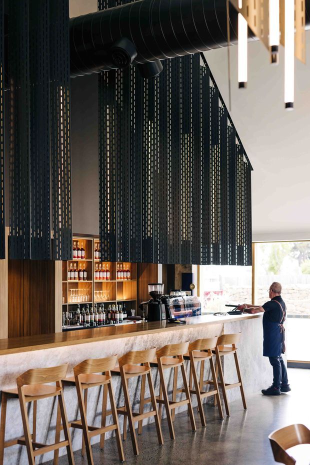 A contemporary commercial bar or café interior with a long stone-faced counter and timber bar top, lined with wooden stools. Behind the counter are built-in dark timber display shelves stocked with bottles and glassware. A dramatic suspended black metal slatted screen conceals exposed ductwork overhead, while slim linear pendant lights hang above. The space has polished concrete flooring, expansive glazing, and a warm yet industrial material palette.