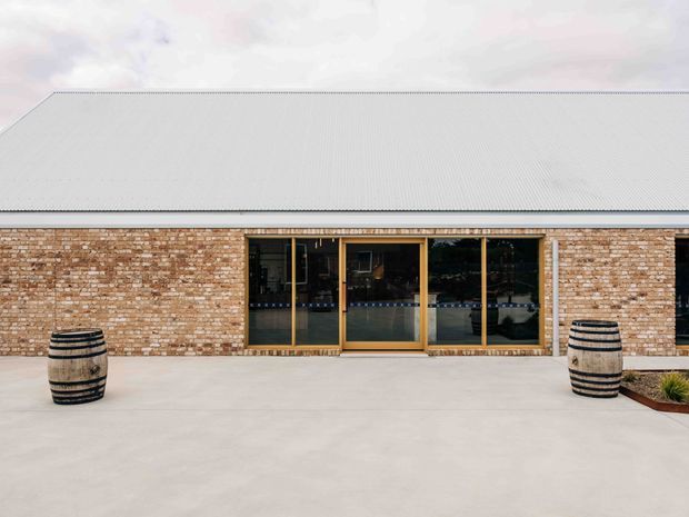 Front-facing exterior view of a low, single-storey brick building with a broad corrugated metal gable roof, large timber-framed glazed doors or windows, and a wide pale concrete forecourt. Two decorative wooden barrels flank the entrance, with minimal landscaping at the right edge.