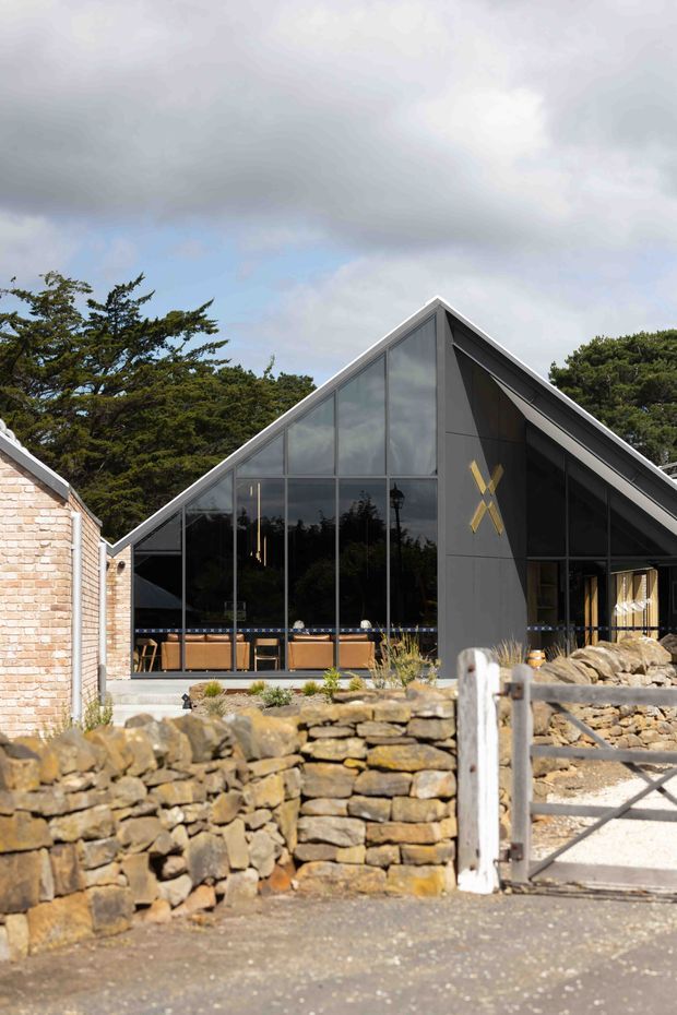 Exterior view of a contemporary gable-roof building with a dramatic fully glazed triangular frontage, dark vertical cladding, and a prominent yellow geometric emblem. A pale brick structure adjoins it on the left. In front are dry-stone walls, gravel paving, and a simple timber gate, with mature trees behind and an overcast sky.