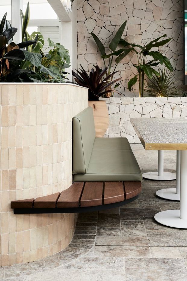 A stylish commercial or hospitality interior featuring a curved built-in banquette wrapping around a rounded tiled partition. The seating combines muted sage-green upholstery with a dark timber platform and curved timber slats. Behind it is a pale irregular stone-clad wall with abundant tropical indoor planting. Small round tables with light terrazzo-like tops and white pedestal bases occupy the right side. The floor is finished in large mottled grey-beige stone-look tiles, and bright glazing creates an airy atmosphere.