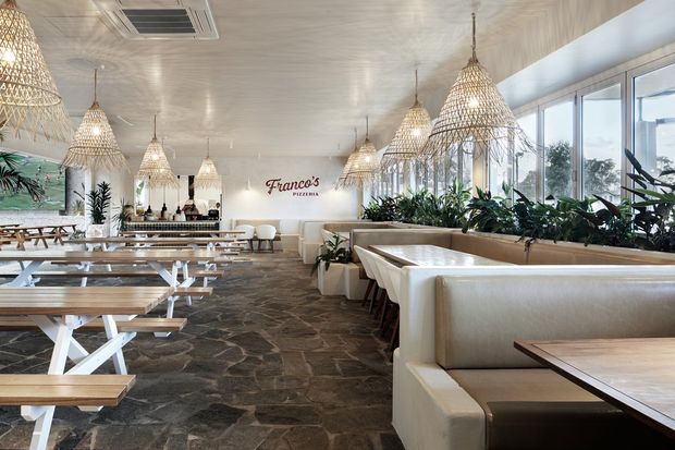 A bright, spacious pizzeria dining room with long communal timber tables and white trestle-style bases, upholstered booth seating, abundant indoor plants, large windows, and numerous woven rattan pendant lights. The interior features a textured light ceiling, dark irregular stone-look flooring, and a prominent wall sign reading “Franco’s Pizzeria.”