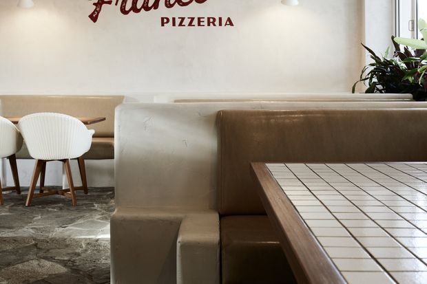 Interior view of a pizzeria dining area featuring built-in upholstered booth seating, a tiled tabletop with timber edging, white molded dining chairs, and a textured stone-look floor. A partial red pizzeria sign and indoor planting are visible against the light wall.
