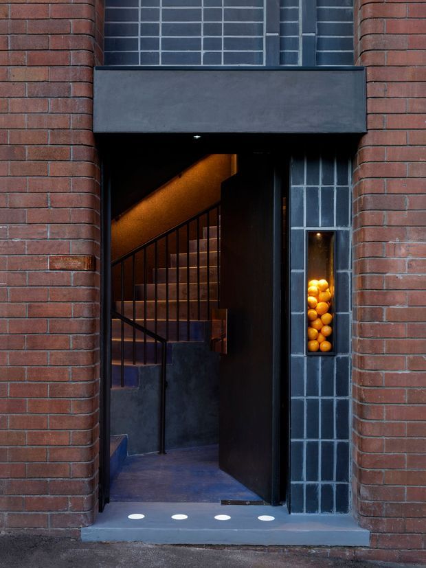 A close-up view of a dark, contemporary brick building entrance. Red brick walls frame a black metal and timber doorway, with a narrow blue-grey tiled surround and inset display niche filled with oranges. Through the open door, a warmly lit stairway with black metal balustrades is visible. The entry threshold includes small circular white floor details, creating a moody and distinctive facade composition.