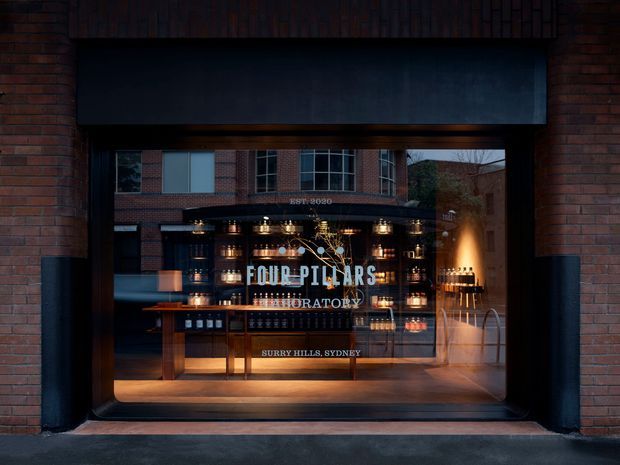 A dark, polished storefront facade in brick with a large glazed opening displaying a warmly lit Four Pillars gin laboratory and bar interior. The entrance features black metal framing, timber display cabinetry, illuminated bottles and shelving, and prominent branding on the glass. The image has a moody, refined commercial hospitality character.