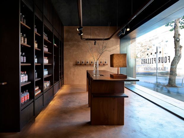 A dark, refined retail interior viewed from inside a boutique shop. Full-height black shelving on the left displays bottles and products, while a central dark timber display counter and a warm textured table lamp occupy the space. The rear wall has warm brown plaster or concrete finishes with minimal shelving and branch-like decor. Large floor-to-ceiling glazing on the right opens the shop to the street, creating strong contrast between the moody interior and bright exterior. Track lighting, polished concrete flooring, and dark ceilings contribute to an industrial yet sophisticated atmosphere.